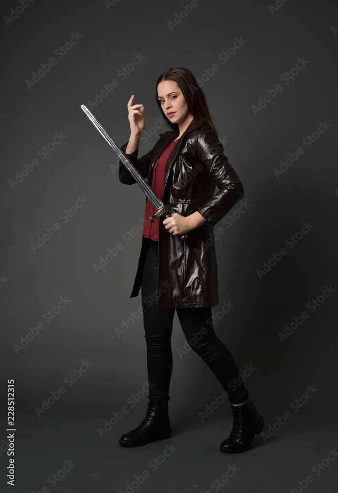 full length portrait of brunette girl wearing long leather coat and boots. standing pose and holding a sword on grey studio background. full length portrait of brunette girl wearing long leather coat and boots. standing pose and holding a sword on grey studio background.