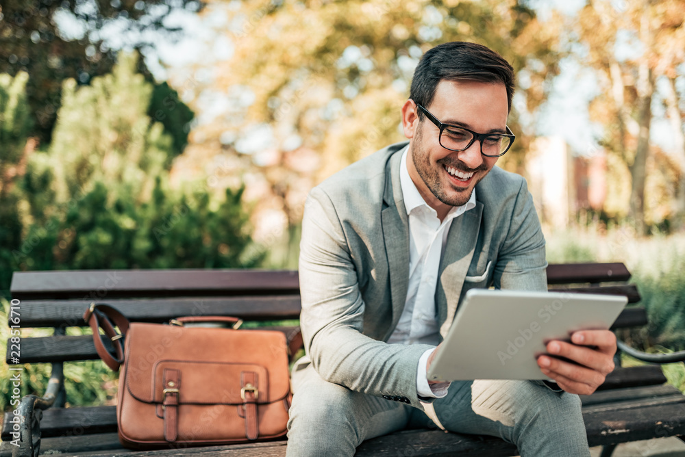 Smiling business man using tablet while sitting on the bench. - obrazy, fototapety, plakaty 