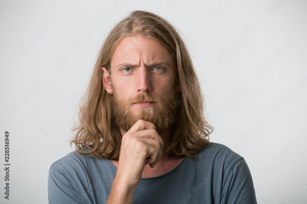 Young guy with a beard, mustache, blond hair to the shoulders and blue eyes  looks contemplative and thoughtful, focused, eyebrows frowned. Keeps a hand  on the chin, isolated over white background Stock
