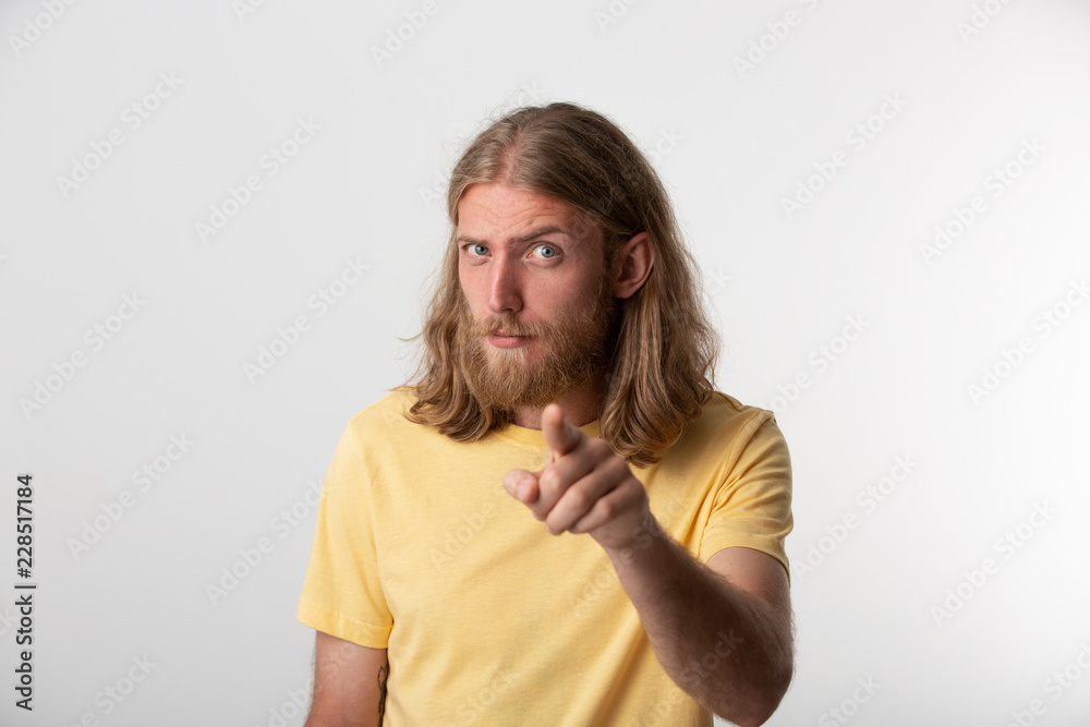 Hey you! Closeup photo of flirty young guy with a beard, mustache, blond hair to the shoulders and blue eyes looking at camera and pointing finger isolated over white background Hey you! Closeup photo of flirty young guy with a beard, mustache, blond hair to the shoulders and blue eyes looking at camera and pointing finger isolated over white background