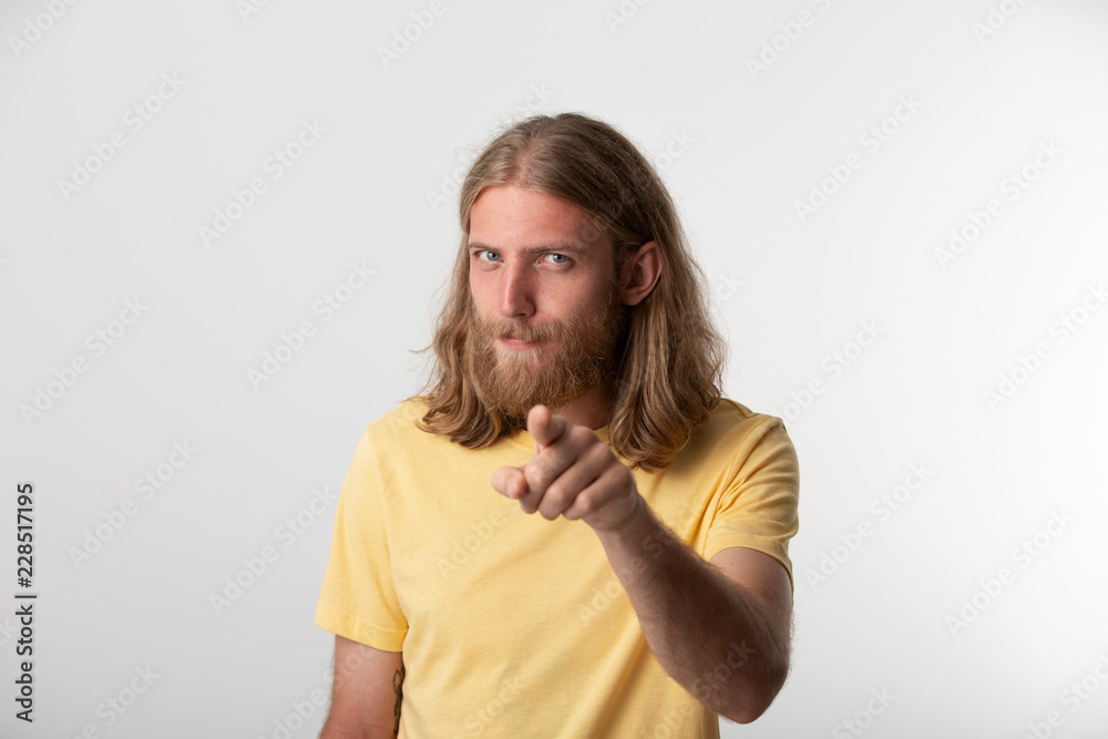 Hey you! Closeup photo of flirty young guy with a beard, mustache, blond hair to the shoulders and blue eyes looking at camera and pointing finger isolated over white background Hey you! Closeup photo of flirty young guy with a beard, mustache, blond hair to the shoulders and blue eyes looking at camera and pointing finger isolated over white background