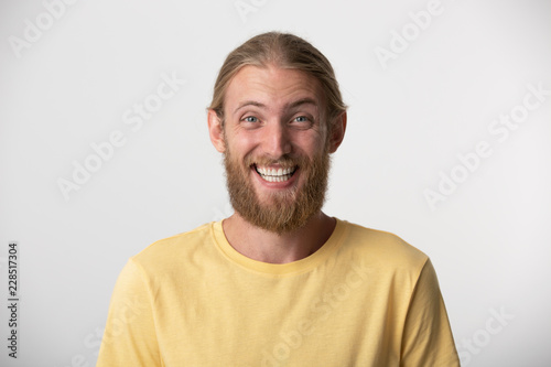 Cheerful young man with beard and blond hair put back smiles happily, has excited expression, dresssed casually, celebrates his anniversary or promotion at work, isolated over white studio background.