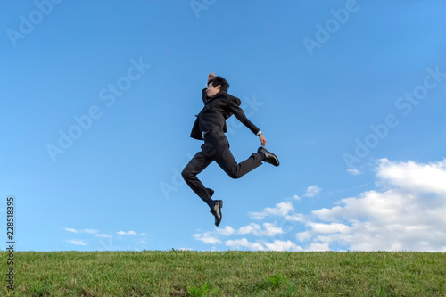 青空をバックにジャンプするスーツ姿の若いビジネスマン1人。元気・パワー・喜び・挑戦イメージ