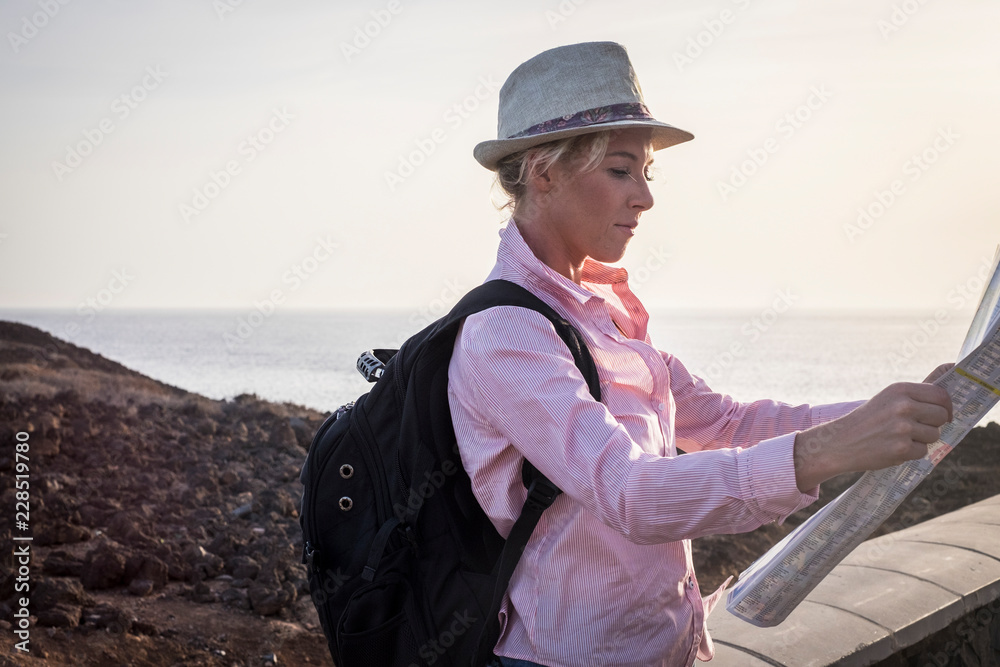 young blonde female traveler with black backpack read the map to choose the next destination. Coast and ocean in background. Outdoor travel concept