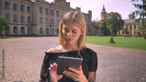 Pretty caucasian short-haired blonde woman is using her tablet and standing still on the sunndy urban street photo