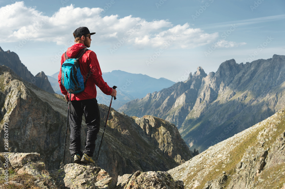A bearded man in sunglasses and a cap with a backpack stands on top of a rock and looks into a rocky valley high in the mountains. The concept of tourism and easy trekking in the mountains outdoor