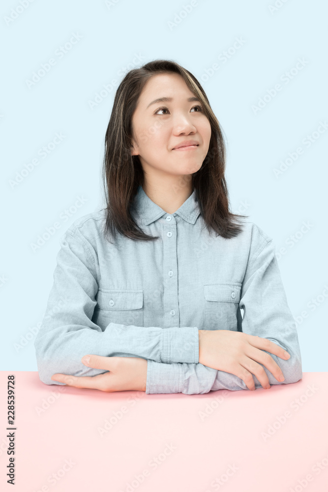 Serious business woman sitting at table, looking up isolated on trendy blue studio background. Beautiful, young face. Female half-length portrait. Human emotions, facial expression concept. Serious business woman sitting at table, looking up isolated on trendy blue studio background. Beautiful, young face. Female half-length portrait. Human emotions, facial expression concept.