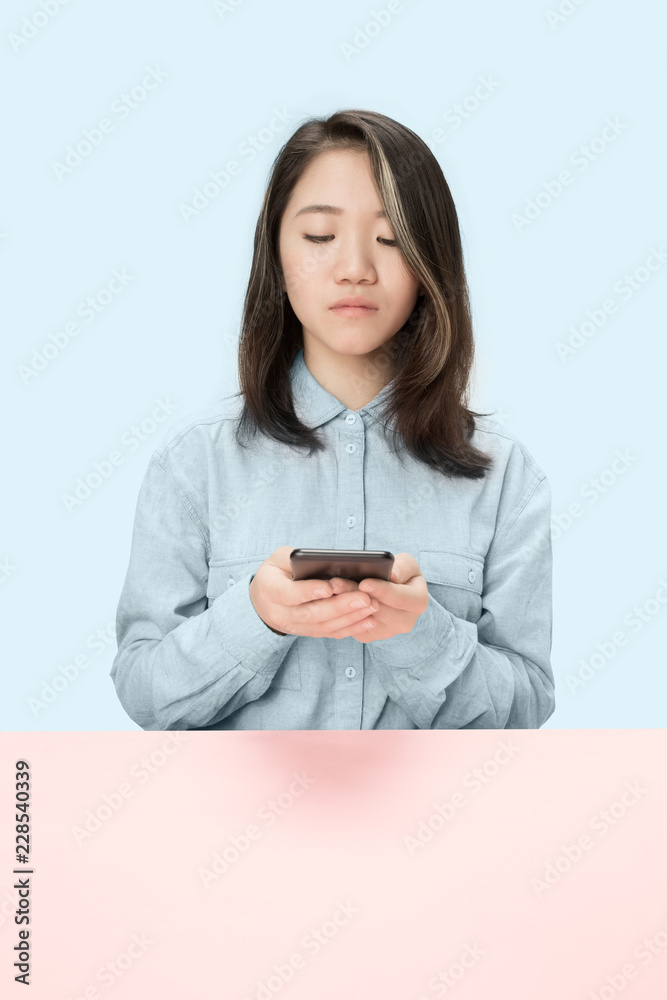 Serious business woman sitting at table, looking at phone isolated on trendy blue studio background with mobile phone. Beautiful, young face. Female half-length portrait. Human emotions, facial Serious business woman sitting at table, looking at phone isolated on trendy blue studio background with mobile phone. Beautiful, young face. Female half-length portrait. Human emotions, facial