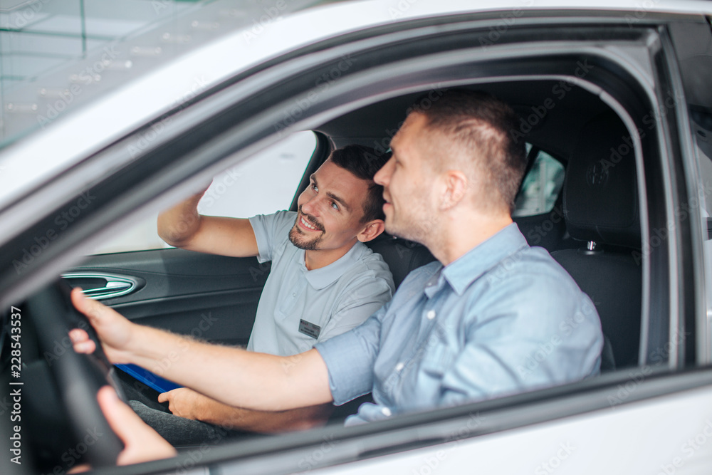 Nice and positive young men sit in car and look up. Bearded guy points. They sit on front sits. Car is white. Young men smile.