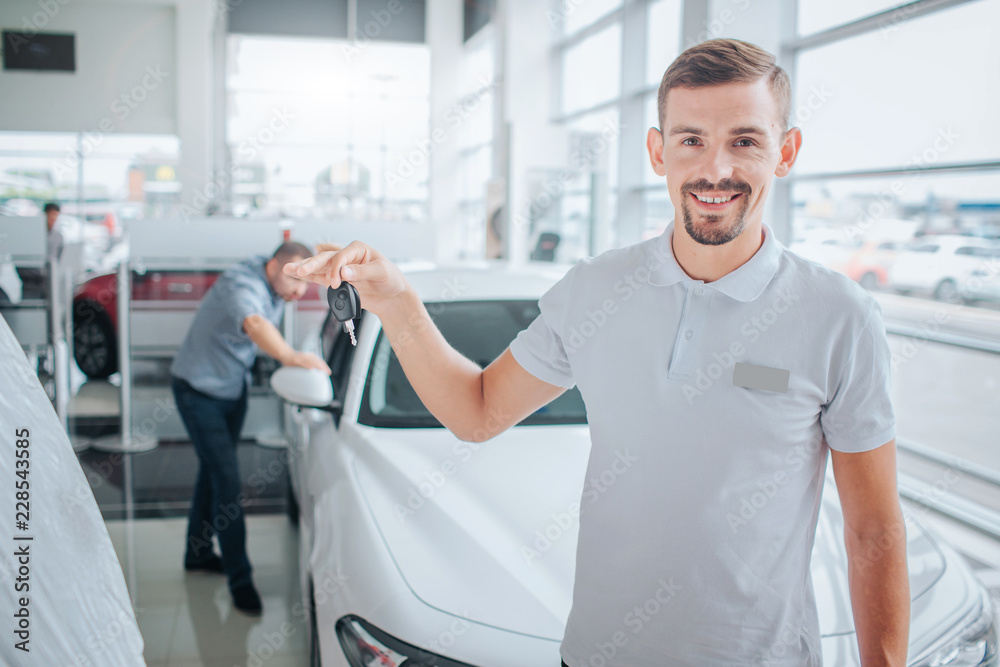 Obraz premium Picture of car deeler stands and looks on camera. He smiles and holds car key. Potential purchaser stands and leans on white car. He looks inside of it. with seriousness. They are in dealing store.