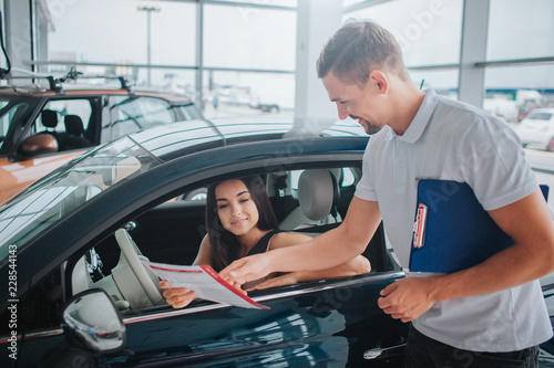 Nice consultant stands at black car and points on paper. He holds plastic tablet in other hand. Nice and young woman sits in car and looks at document. She is concentrated.