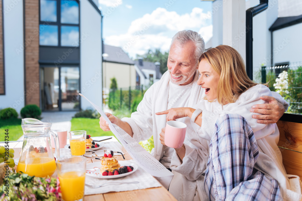 Surprising news. Beautiful blonde-haired woman feeling surprised while reading morning news sitting near her bearded husband Surprising news. Beautiful blonde-haired woman feeling surprised while reading morning news sitting near her bearded husband