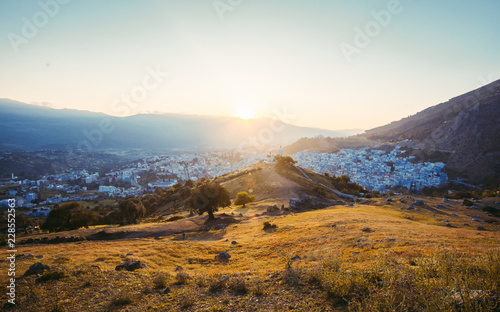 Adventurous man sitting on top of the mountain enjoying the beautiful view during a vibrant sunset. Chefchaouen panorama, blue city skyline on the hill, Morocco © Pavel Kašák