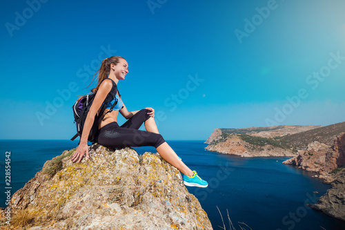 Young hikers woman sitting on a rock and looking at sea and mountain landscape. Happy athlete relaxes on a stone and enjoying the view from the cliff.
