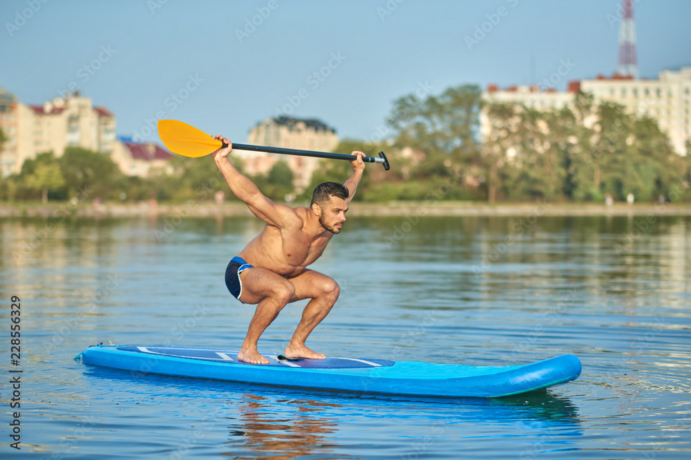 Sportsman holding long oar over head, swimming on paddle board in city lake.