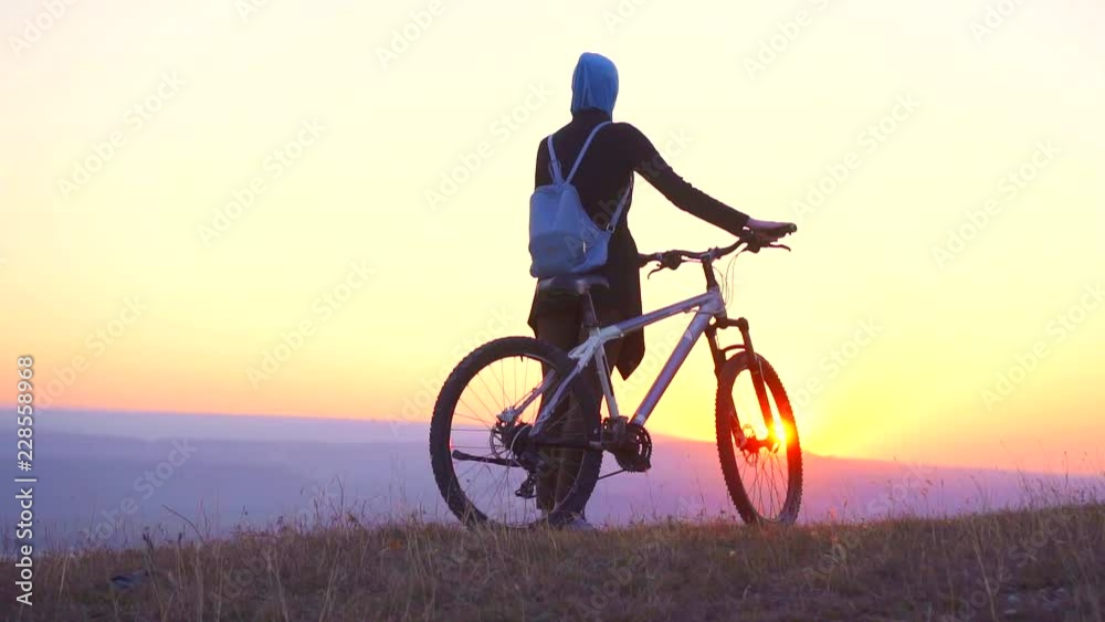 Muslim woman in a hijab with a backpack with a bicycle from the mountain looks at a sunset,slow mo Muslim woman in a hijab with a backpack with a bicycle from the mountain looks at a sunset,slow mo