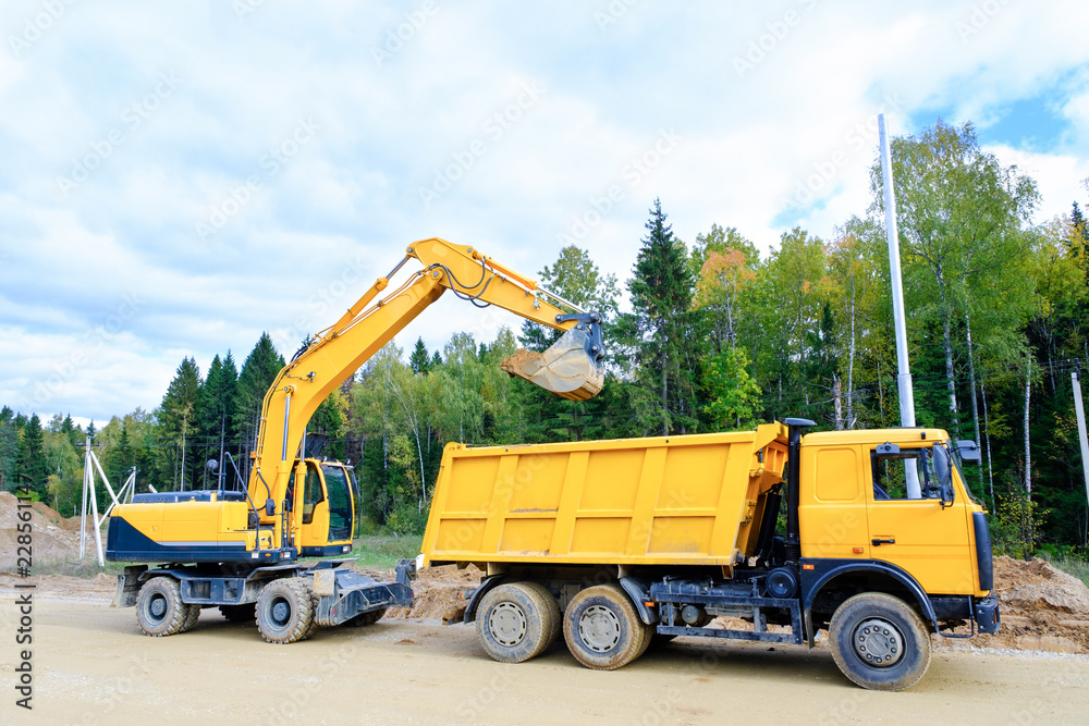 The wheel excavator loads the earth with a bucket to the body of a multi-ton dump truck on the construction site The wheel excavator loads the earth with a bucket to the body of a multi-ton dump truck on the construction site