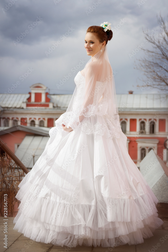 Fototapeta premium Portrait of a bride in a wedding dress and with a long veil on the background of the ancient city and a cloud in the autumn day.