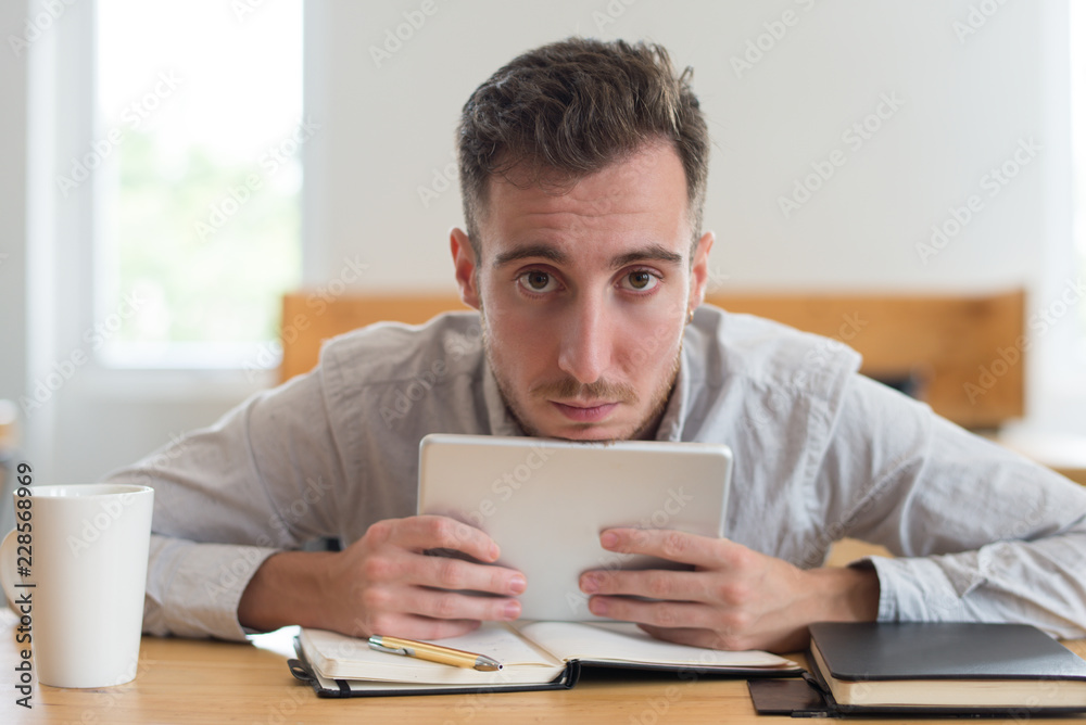 Serious male student holding tablet and looking at camera in classroom. Young man browsing on gadget at desk. Education and technology concept. Front view.