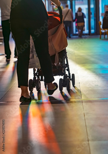 Young mother walking with baby carriage in the shopping center, from the back, evening sun shine reach inside through the swing door.