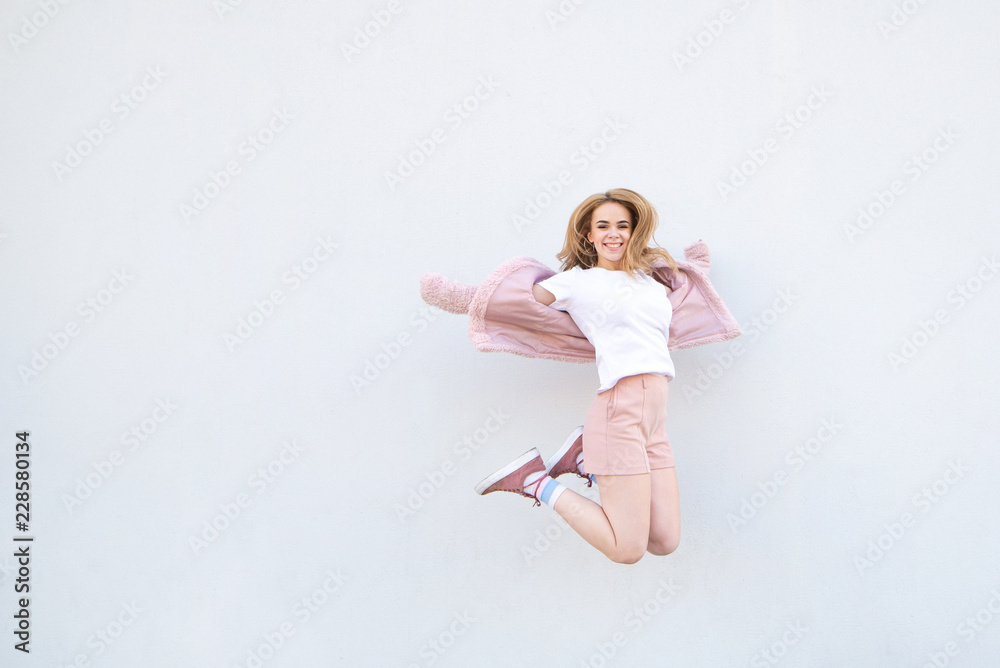 Young blonde girl in pink clothes jumping against the background of a white wall and smiling. Attractive happy girl levitation on the background of a white wall. Copyspace Young blonde girl in pink clothes jumping against the background of a white wall and smiling. Attractive happy girl levitation on the background of a white wall. Copyspace