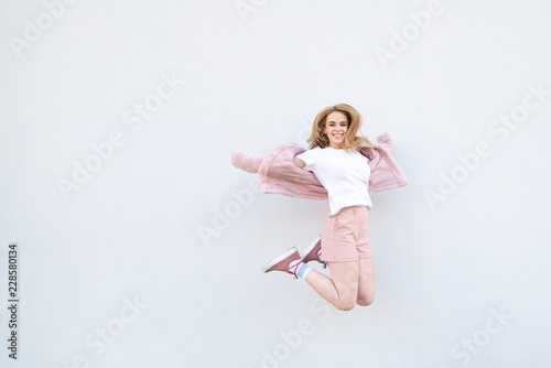 Young blonde girl in pink clothes jumping against the background of a white wall and smiling. Attractive happy girl levitation on the background of a white wall. Copyspace