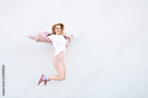 Happy smiling girl in pink stylish clothes in the air on a white background. Attractive girl in a pink coat and shorts jumping against the background of a white wall. Copyspace