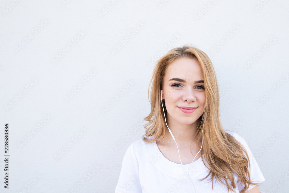 Portrait of a blond girl in a white T-shirt on a white background, listens to music in the headphones, looks into the camera and smiles. Copyspace Portrait of a blond girl in a white T-shirt on a white background, listens to music in the headphones, looks into the camera and smiles. Copyspace