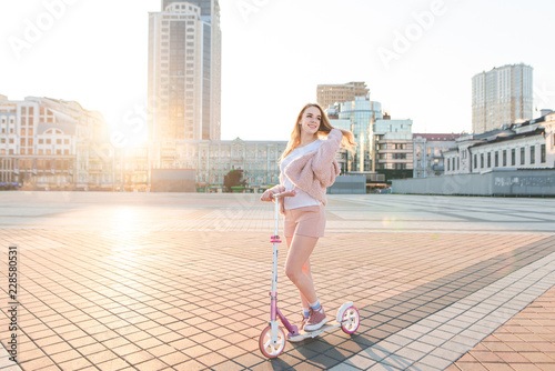 Happy model in pink dress and pink kick scooter pose against the background of the city landscape in the sunset and smiles. Cute girl walking on a scooter.