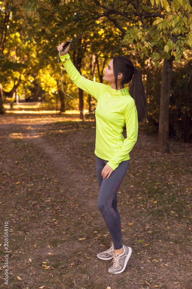 Beautiful young girl in sportswear takes a selfie on the phone and listens to music while jogging in the forest, park in the fall at sunset. Sports lifestyle