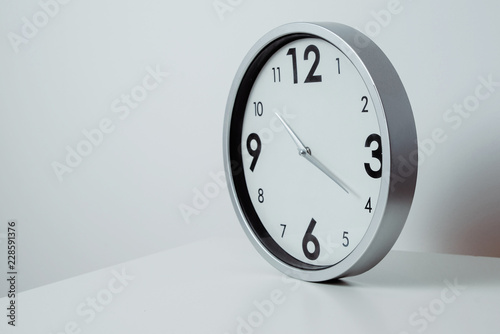 A view of a silver, large wall clock. Side view of a clock showing the time lying on a white table. The concept of passing time, ending work, returning to school and work tasks.