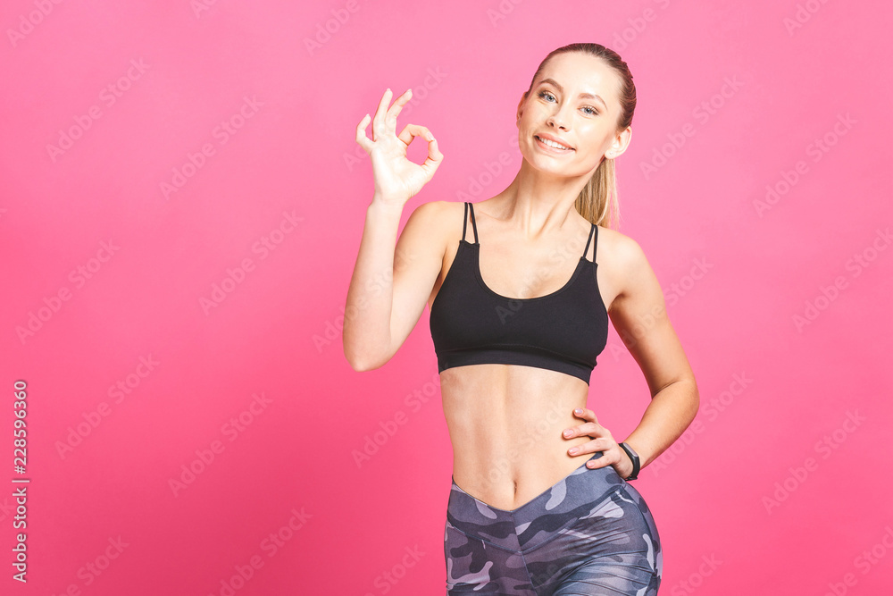 Beautiful young woman in sports wear showing Ok sign, looking at camera and smiling, isolated over pink background. Beautiful young woman in sports wear showing Ok sign, looking at camera and smiling, isolated over pink background.