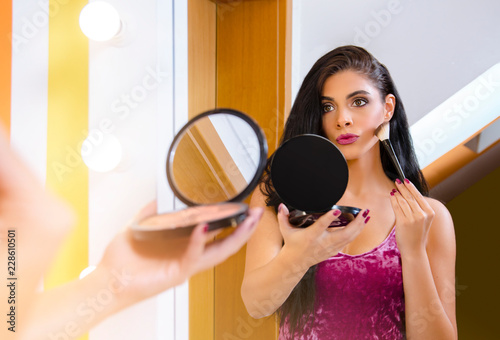 Gorgeous young woman applying makeup with brush, looking herself into the mirror 
