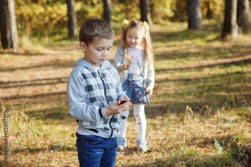 Brother and sister in the autumn forest. A boy in a plaid shirt carefully works with a smarphone.