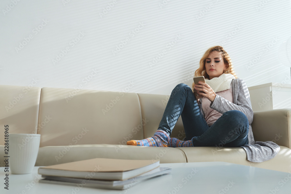 Attractive young in scarf and warm clothes resting on sofa and texting friends Attractive young in scarf and warm clothes resting on sofa and texting friends