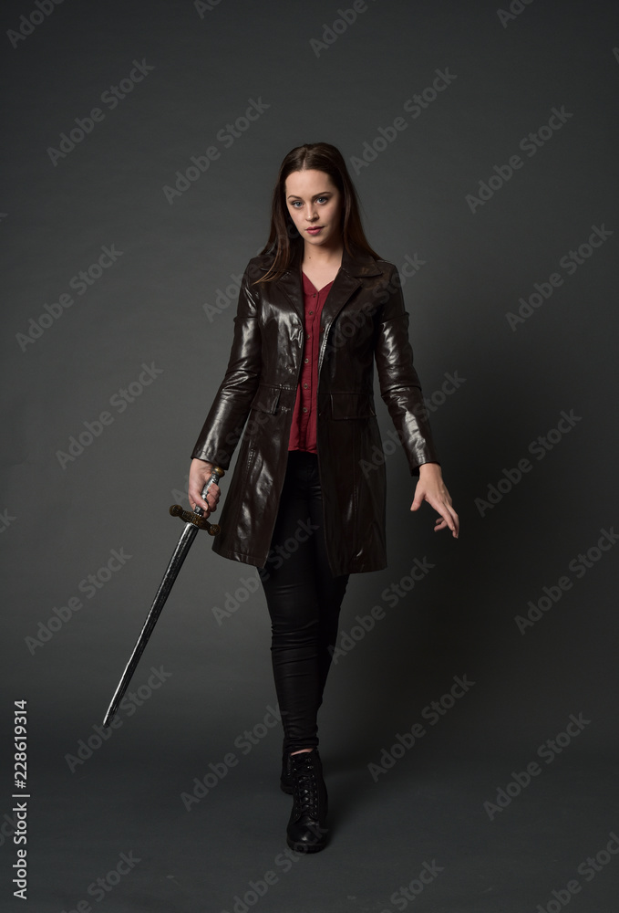 full length portrait of brunette girl wearing long leather coat and boots. standing pose and holding a sword on grey studio background. full length portrait of brunette girl wearing long leather coat and boots. standing pose and holding a sword on grey studio background.