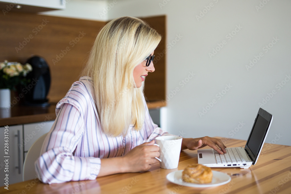 blond woman in pajamas drinking coffee and using laptop in the kitchen