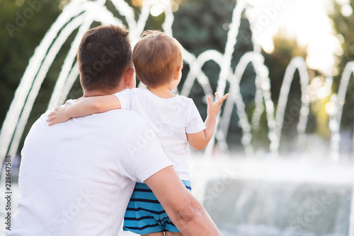 Little child embracing his father while both are watching at fountain water strings, view from the back. Family vacation concept. Room for text #228647586