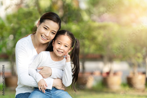 Portrait of young happy asian mother and little cute daughter smiling, sitting and looking at camera at outdoor public nature park with copy space in family bonding and casual relax lifestyle concept. © ChayTee