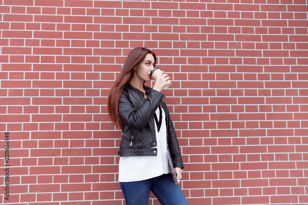Red-haired girl in a black leather jacket holds a laptop and drinks coffee against a red brick wall