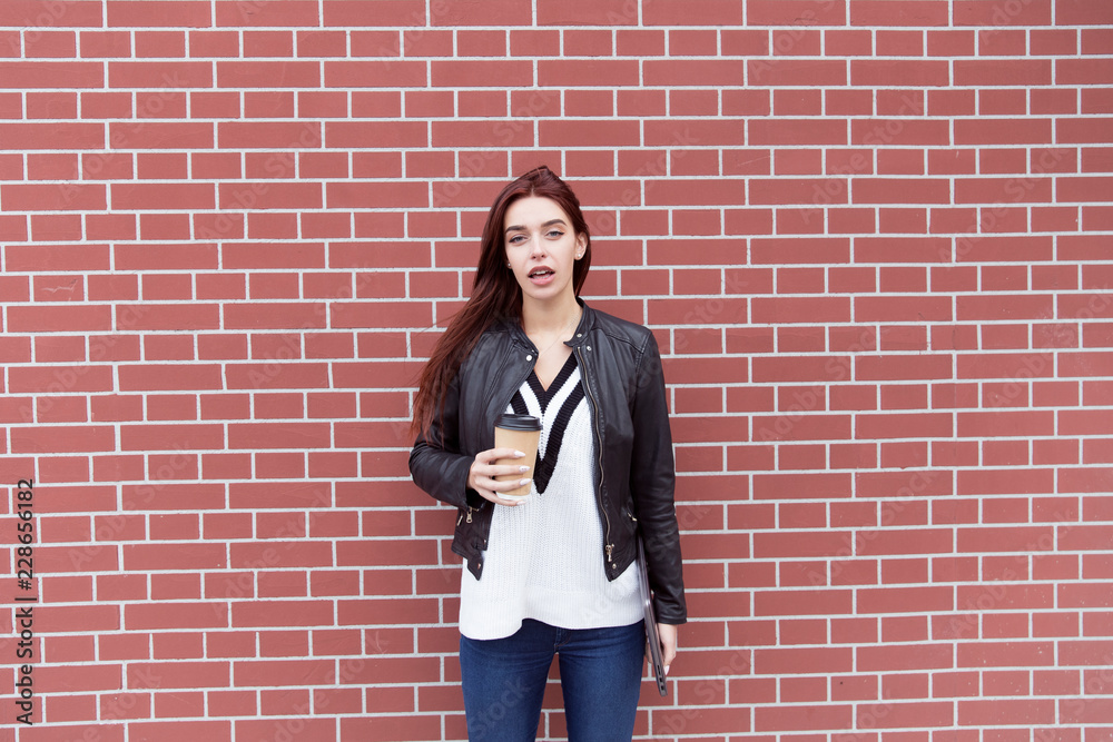 Red-haired girl in a black leather jacket holds a laptop and drinks coffee against a red brick wall Red-haired girl in a black leather jacket holds a laptop and drinks coffee against a red brick wall