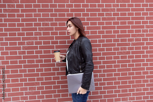 Red-haired girl in a black leather jacket holds a laptop and drinks coffee against a red brick wall