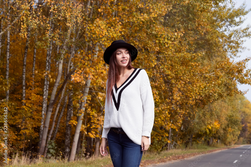 Red-haired girl in a white sweater and a black hat is on the road in the autumn forest