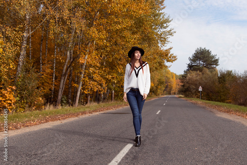 Red-haired girl in a white sweater and a black hat is on the road in the autumn forest