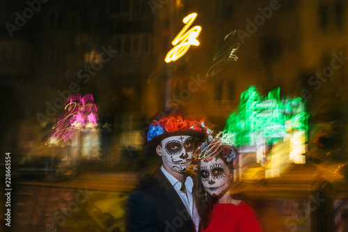 Blur. A man and a woman with a visage, a pattern of skulls, a zombie on the face. Dead at night. A pair of zombies in the dark outside, blurry lights, ghosts. The day of the dead and Halloween.