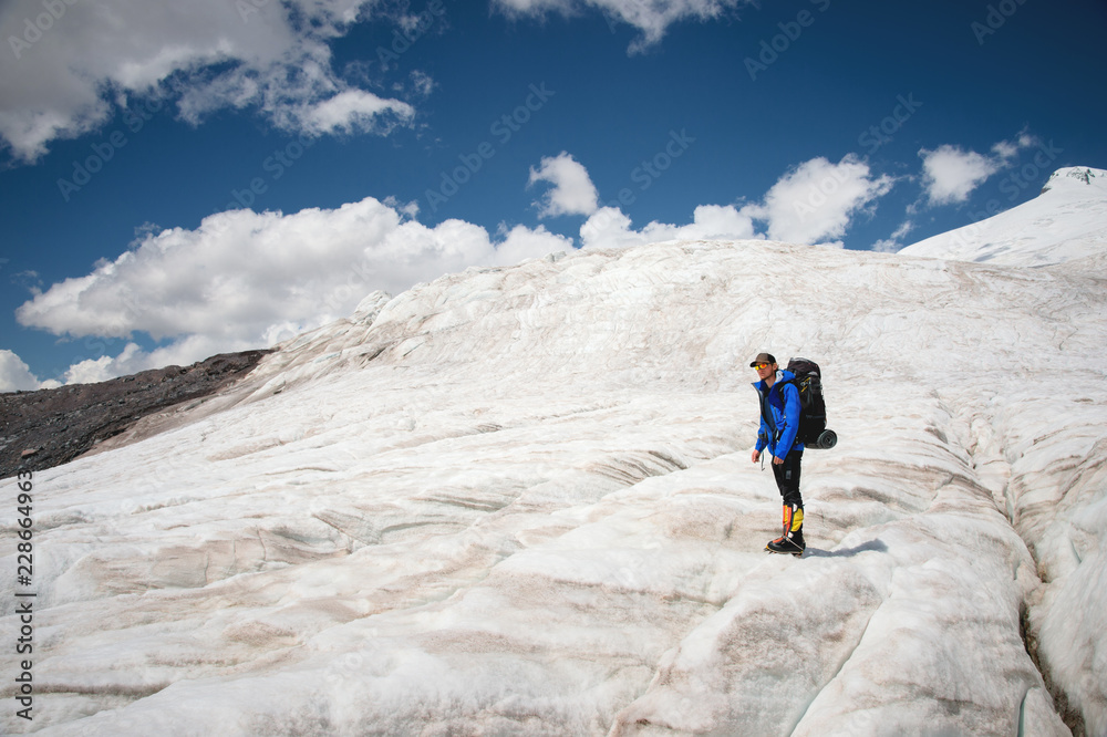 Traveler in a cap and sunglasses with a backpack on his shoulders in the snowy mountains on the glacier against the sky and clouds. Traveler in a natural environment