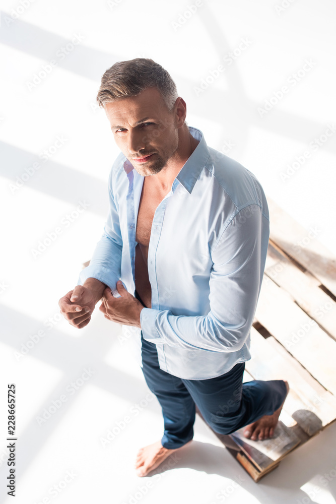 Naklejka premium high angle view of handsome adult man buttoning shirt and looking at camera on white