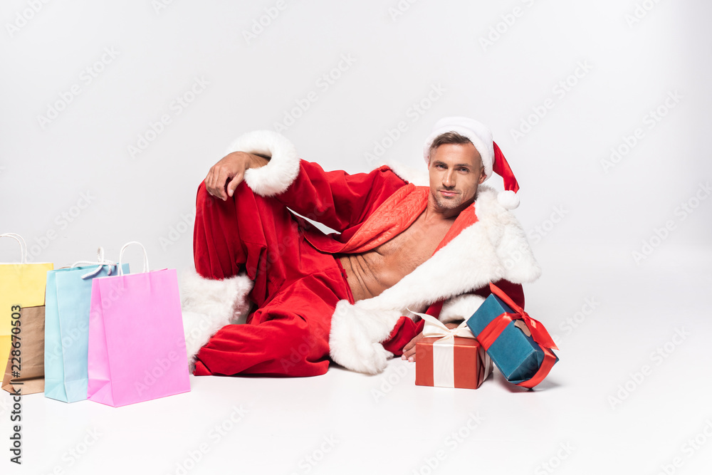 handsome man in santa costume lying with gift boxes and shopping bags on grey handsome man in santa costume lying with gift boxes and shopping bags on grey
