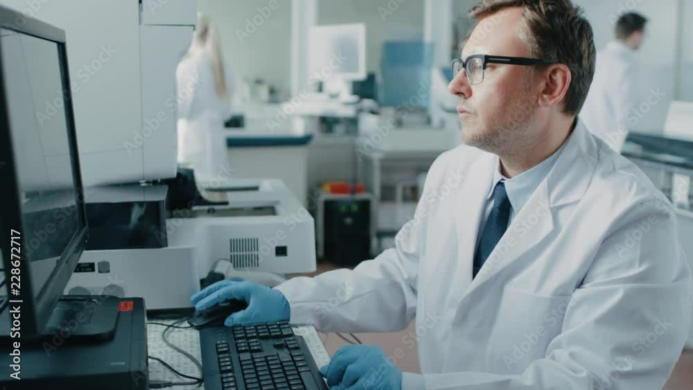 Male Research Scientist Sits at His Workplace in Laboratory, Uses Personal Computer. I the Background Genetics, Pharmaceutical Research Centre with Innovative Equipment. Male Research Scientist Sits at His Workplace in Laboratory, Uses Personal Computer. I the Background Genetics, Pharmaceutical Research Centre with Innovative Equipment.