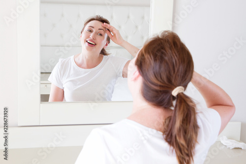 Middle age woman looking in mirror on face wrinkle forehead in bedroom. Wrinkles and anti aging skin care concept. Selective focus